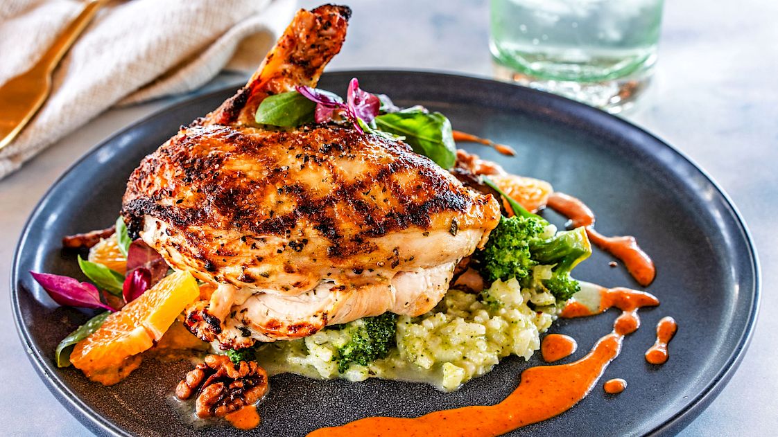 A grilled chicken breast on a bed of mashed vegetables, garnished with broccoli, leafy greens, and drizzled with a red sauce on a black plate.