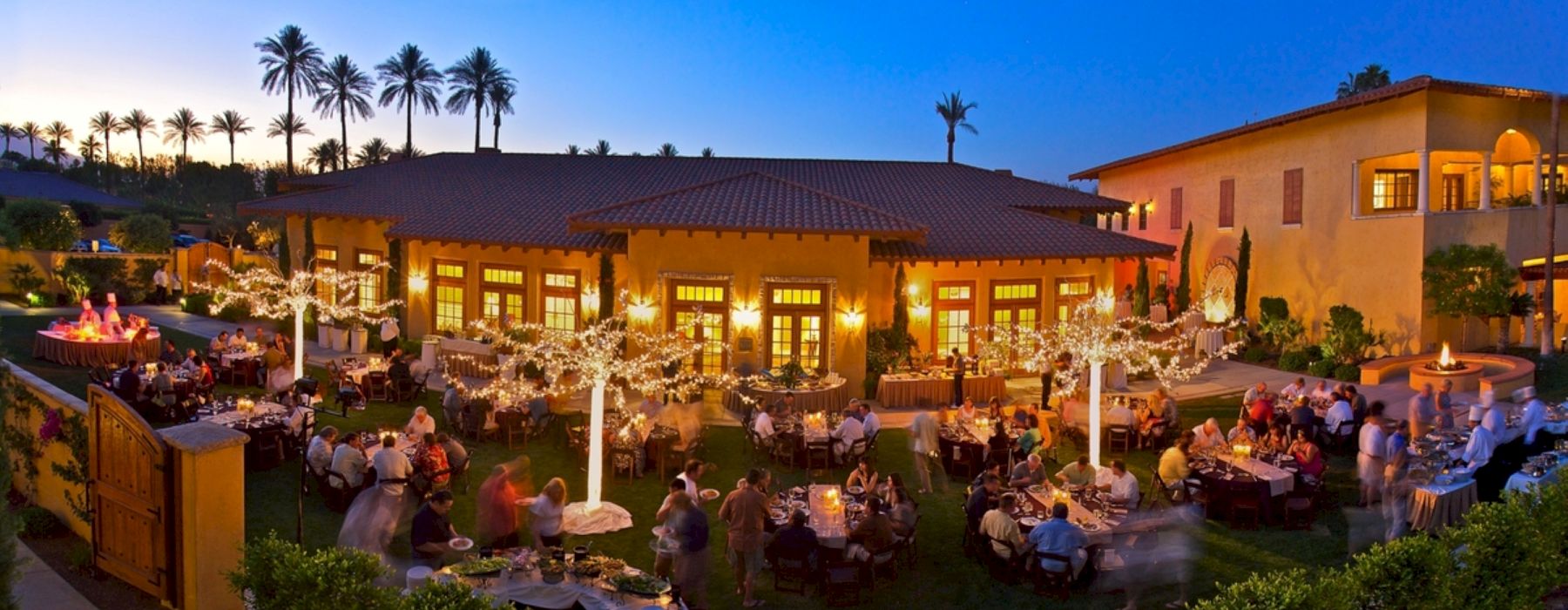 Outdoor event at sunset with attendees seated at tables, surrounded by string lights and palm trees, near a large building with warm lighting.