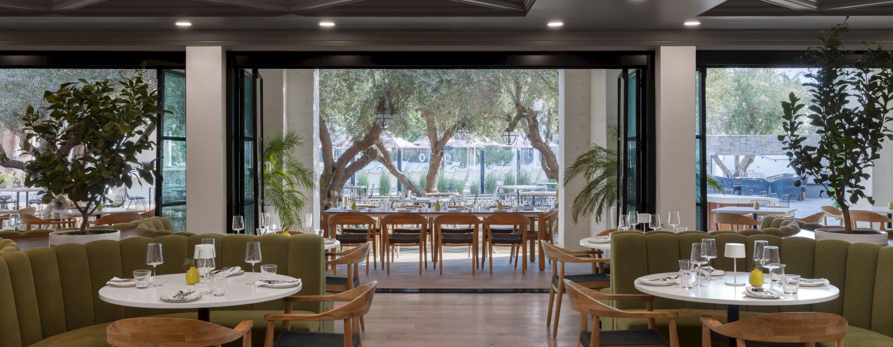 The image shows a modern restaurant interior with neatly set tables, wooden chairs, and green seating. Large windows reveal an outdoor dining area.