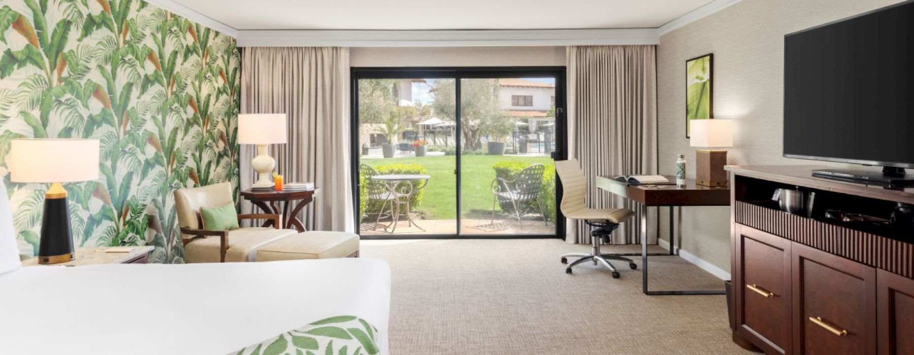 A well-decorated hotel room with a tropical theme features a bed, seating area, large TV, desk, and sliding doors opening to a patio with a garden view.