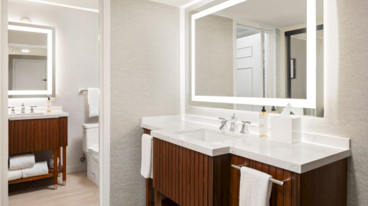A modern bathroom with illuminated mirrors, double sinks, wooden cabinetry, white towels, and a view of the toilet area through an open door.