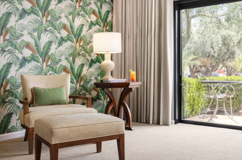 A cozy room with a tropical-themed wall, an armchair, a side table with a lamp, and a sliding door leading to an outdoor patio.