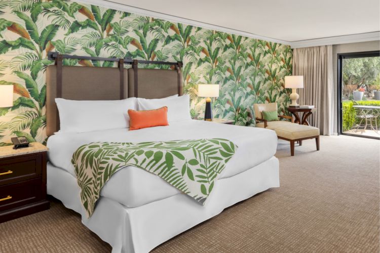 A well-decorated hotel room with tropical-themed wallpaper, a large bed, and an outdoor view through a glass door, ending the sentence.