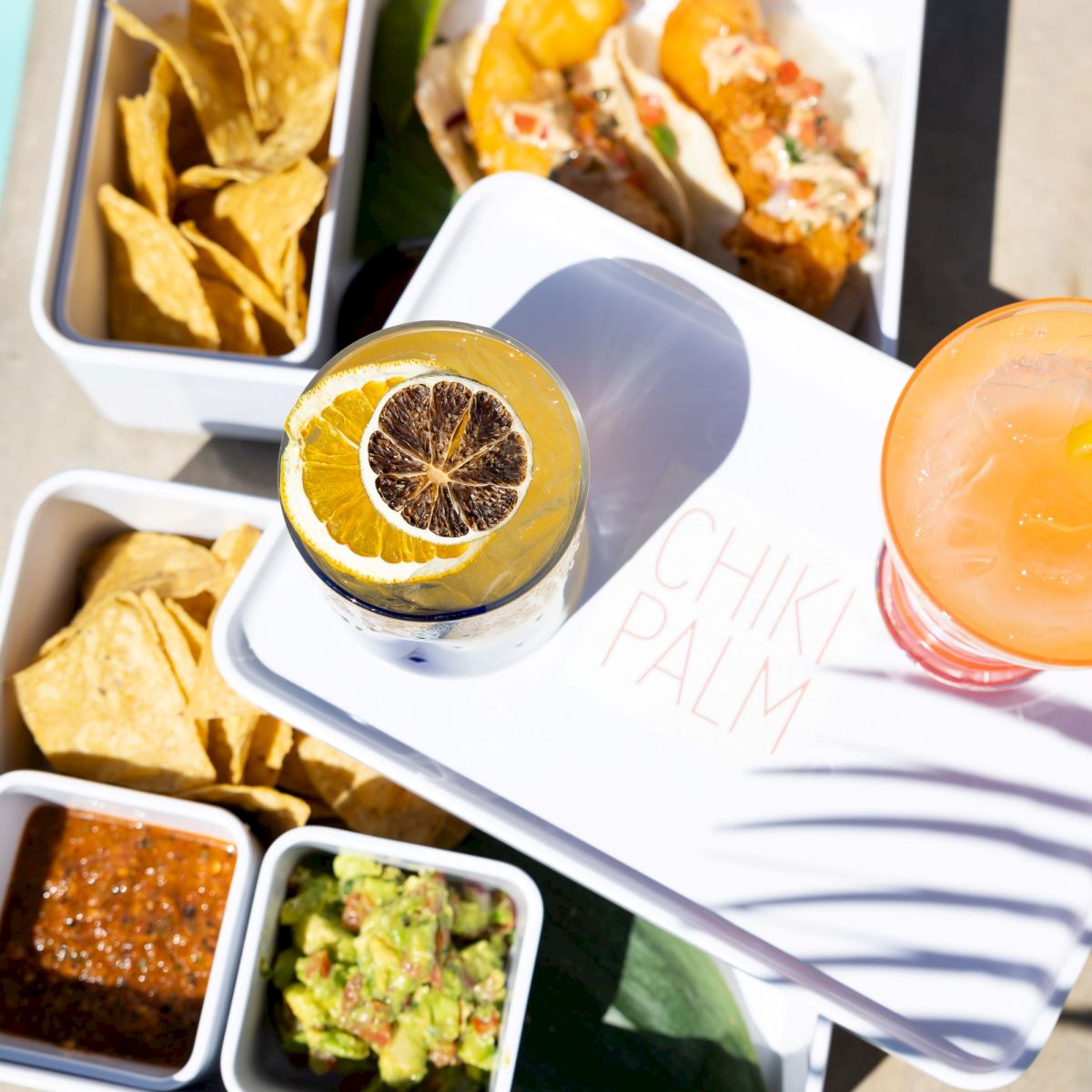 The image shows a poolside table with snacks including tortilla chips, salsa, and guacamole, along with two beverages: one with a lemon slice and one with an orange slice.