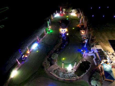Aerial view of a golf course at night, illuminated by colorful lights with visible putting greens, pathways, and surrounding structures.