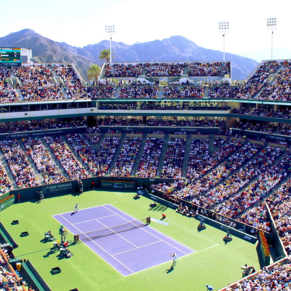A packed stadium hosts a tennis match on a sunny day, with mountains visible in the background and spectators filling the stands.