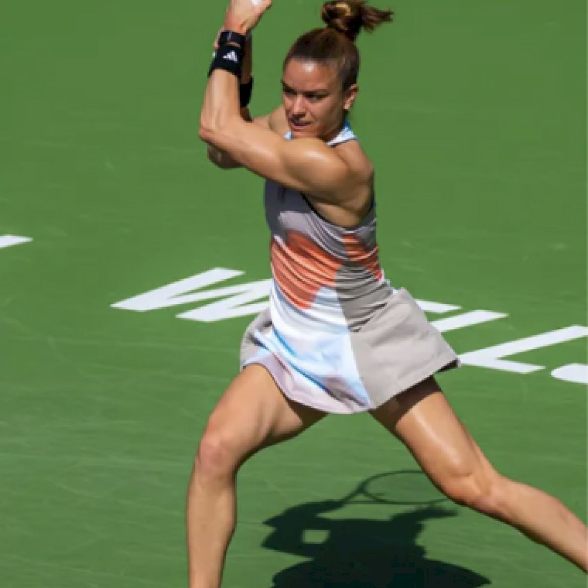 A tennis player, in mid-swing with a racquet, is positioned on a green court, wearing a multicolored outfit and orange shoes.