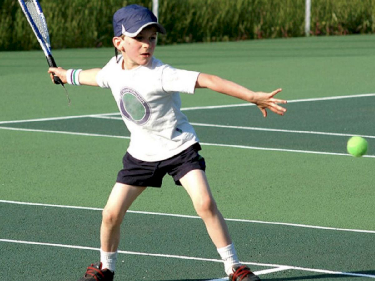 A young child wearing a cap and playing tennis, preparing to hit a tennis ball with a racket on an outdoor court, dressed in athletic attire.
