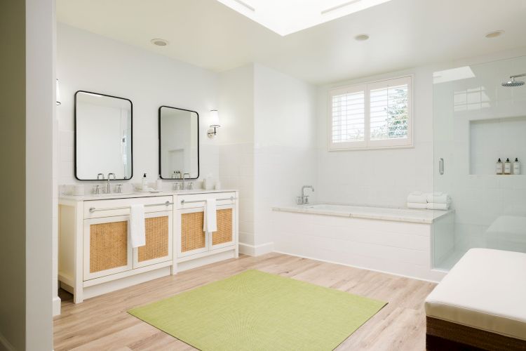 A bright bathroom features dual sinks with mirrors, a bathtub, a shower area, and a green rug on wooden flooring, ending the sentence.