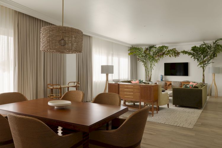 A modern living and dining area features a wooden table, wicker chairs, a TV, large plants, and contemporary decor with neutral tones and ample natural light.