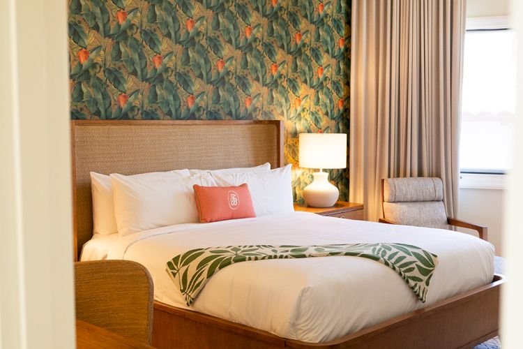 A cozy bedroom with a large bed, patterned wallpaper, wooden furniture, bedside lamp, an armchair, and a window with curtains ending the sentence.