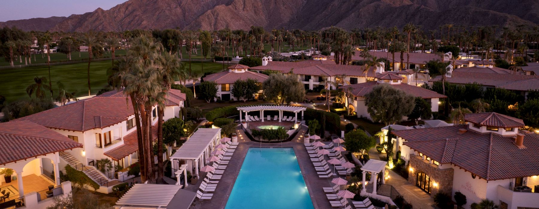 The image shows a luxurious resort with a central swimming pool, surrounded by lounge chairs and buildings, set against a backdrop of mountains.
