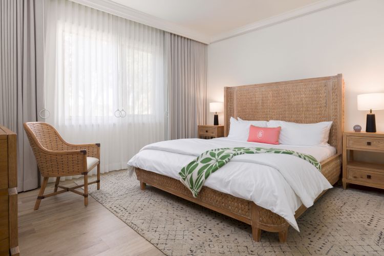 A cozy bedroom with a large bed, two lamps on bedside tables, a wicker chair, a chest of drawers, and a window with light curtains.