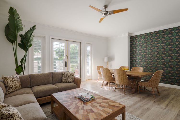 A cozy living room with a sectional sofa, wooden coffee table, dining area, greenery, patterned wallpaper, and large windows with sunlight.
