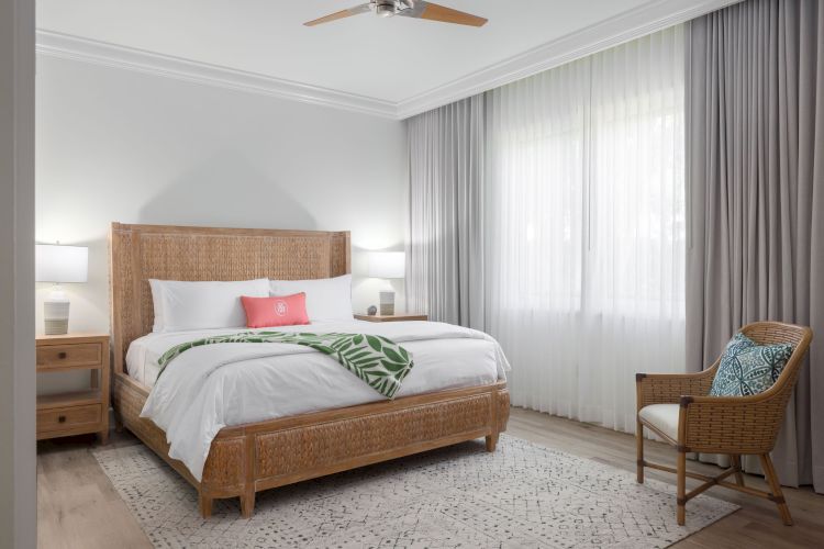 A bright, modern bedroom with a large bed, wicker headboard, bedside tables, lamps, armchair, large window with curtains, and leaf-print blanket on the bed.