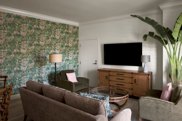 A cozy living room with floral wallpaper, a gray couch, green chairs, a large TV on a wooden console, a glass coffee table, and tall houseplants.