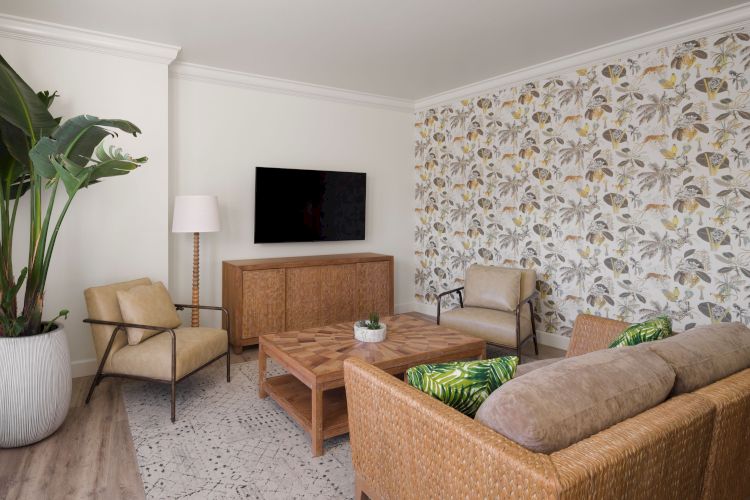 A cozy living room with patterned wallpaper, a wall-mounted TV, wicker furniture, indoor plants, wood coffee table, and lounge chairs in earthy tones.