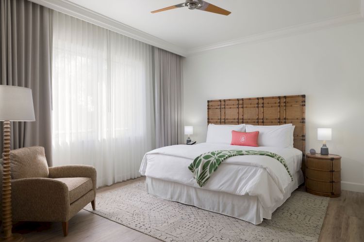 A cozy bedroom with a large bed, two nightstands, a chair, a floor lamp, and a ceiling fan. The decor is neutral with a green throw on the bed.