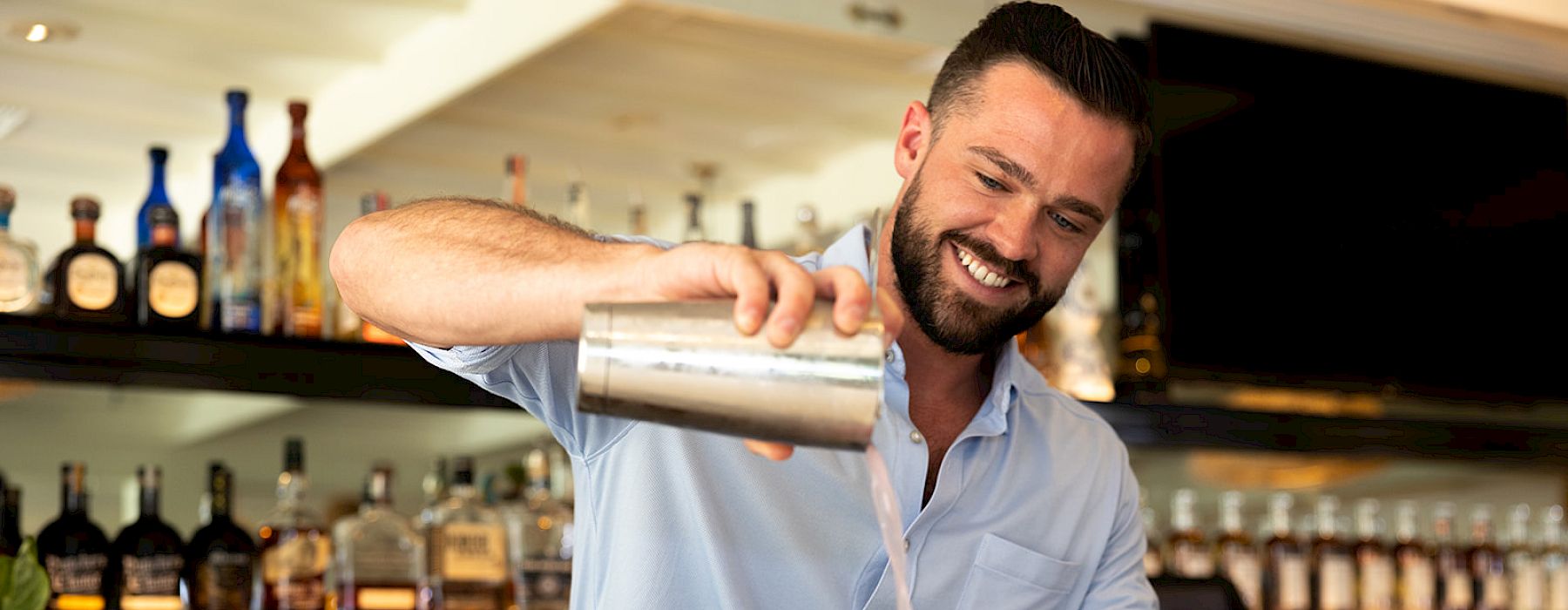 A bartender is pouring a pink cocktail from a shaker into a glass, smiling behind the bar with various bottles in the background.
