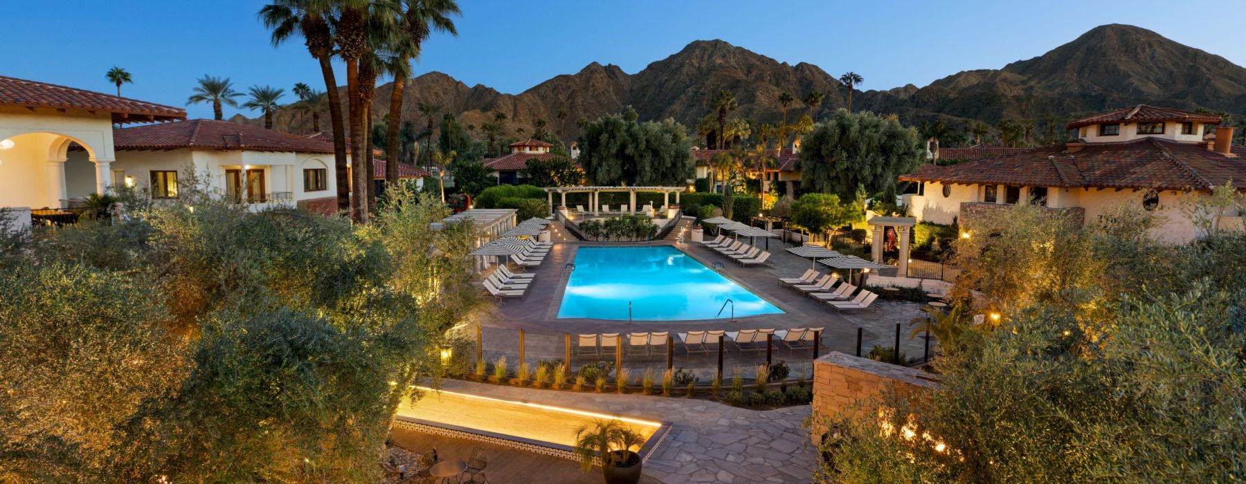 An inviting pool area with sun loungers, surrounded by buildings, lush greenery, and mountains in the background, captured at dusk.
