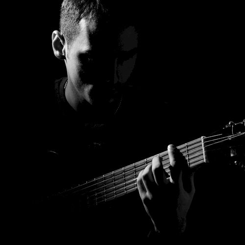 A person is playing an acoustic guitar in a dark setting, with their face and hands partially illuminated by light.