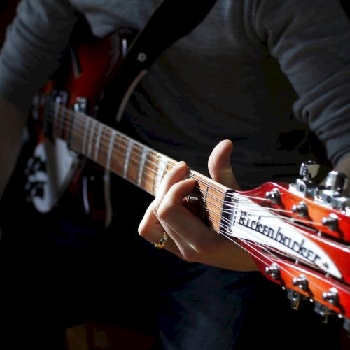 A person is playing a red Rickenbacker electric guitar, focusing on pressing the strings against the fretboard in a dimly-lit setting.