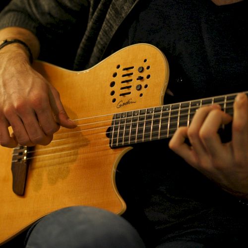A person is playing a wooden acoustic guitar, strumming with one hand while forming chords with the other, wearing casual attire.