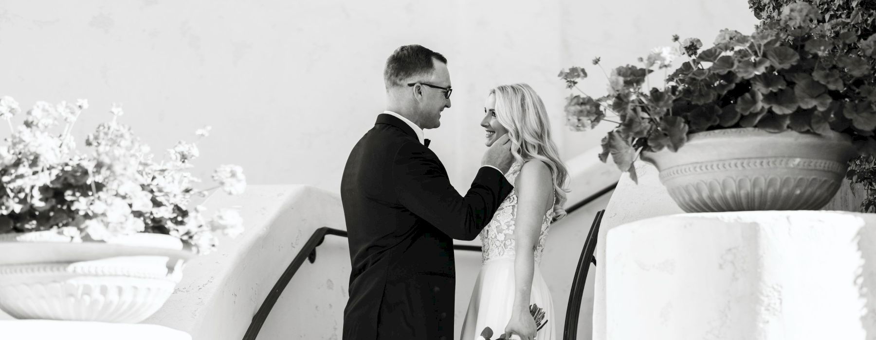 A couple, dressed in wedding attire, stands close on a staircase decorated with flowers and foliage, sharing an intimate moment.