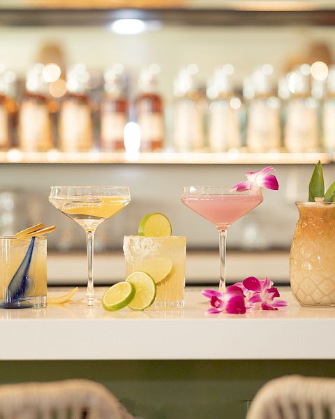 A bar counter with various cocktails and garnishes, including lime slices, orchids, and dried fruits, in front of a selection of bottles.