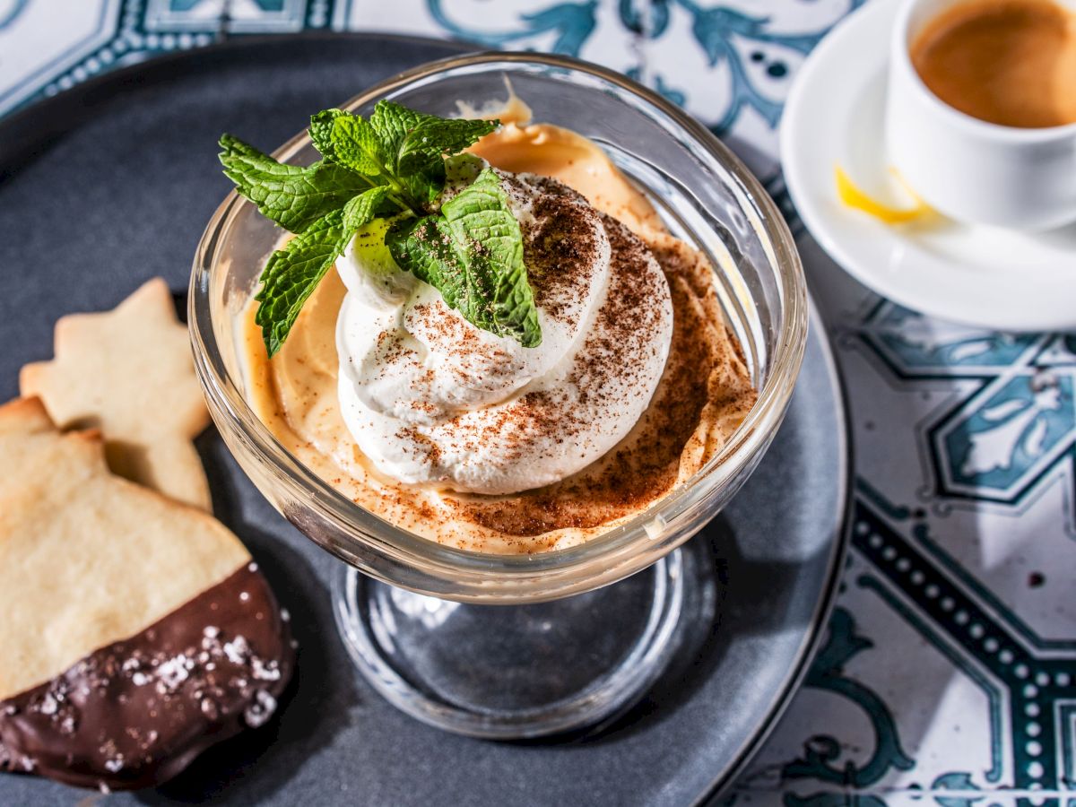 A dessert topped with whipped cream and mint, accompanied by cookies and a cup of espresso on a decorative tablecloth, is shown.