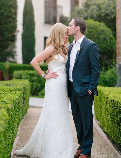 A bride and groom are standing in a garden, with the groom kissing the bride on the forehead while she smiles.