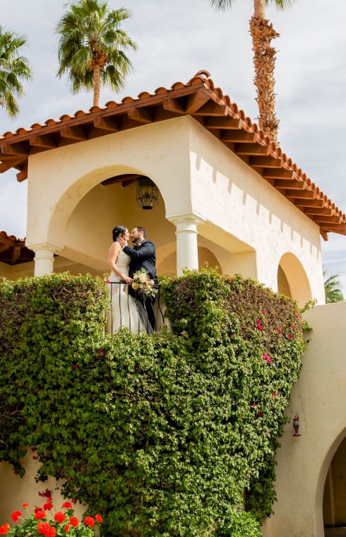 A couple stands on a balcony surrounded by greenery, with palm trees and a Mediterranean-style building in the background.