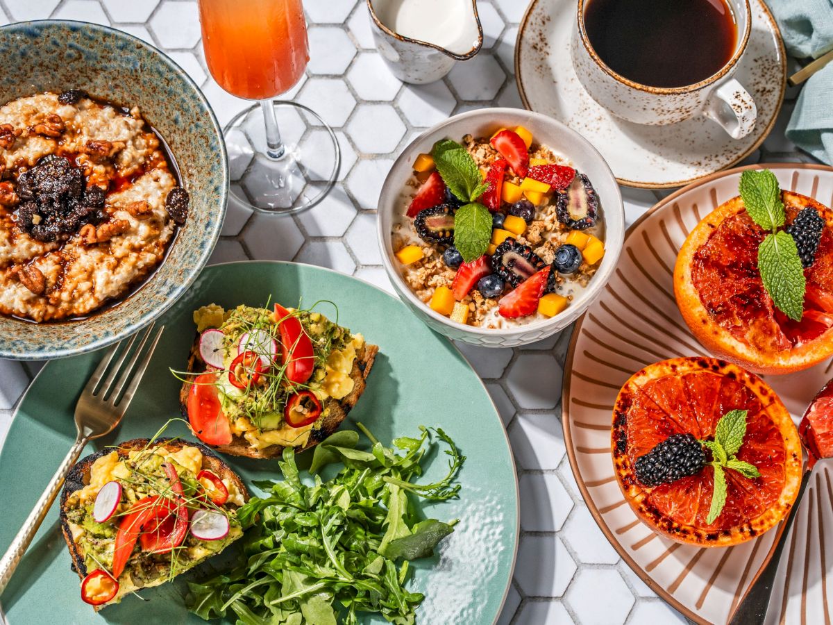 The image shows a breakfast spread with oatmeal, fruit salad, avocado toast, grilled grapefruit, coffee, and a drink, arranged on a white hexagonal tile table.