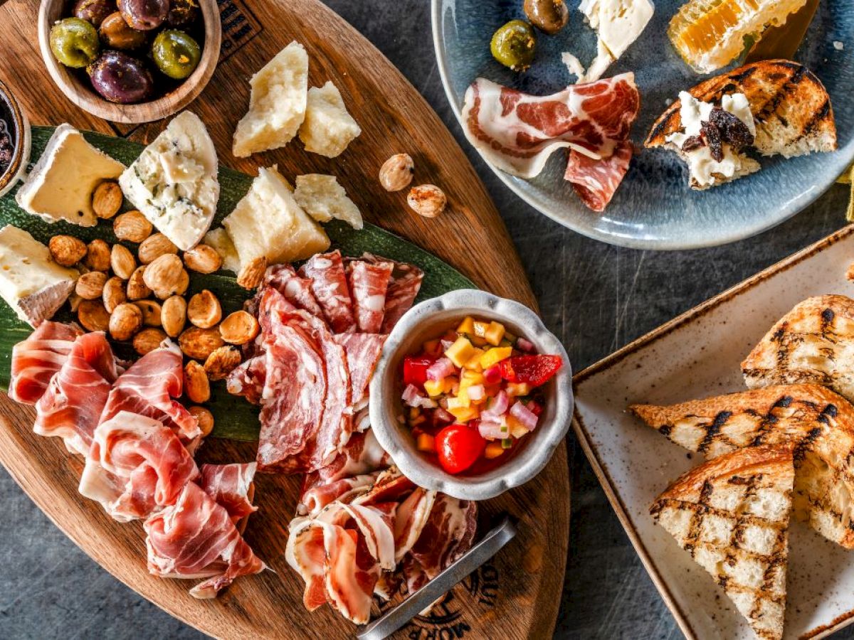 This image shows a charcuterie board with various cheeses, meats, nuts, olives, and a small bowl of mixed vegetables, alongside grilled bread.
