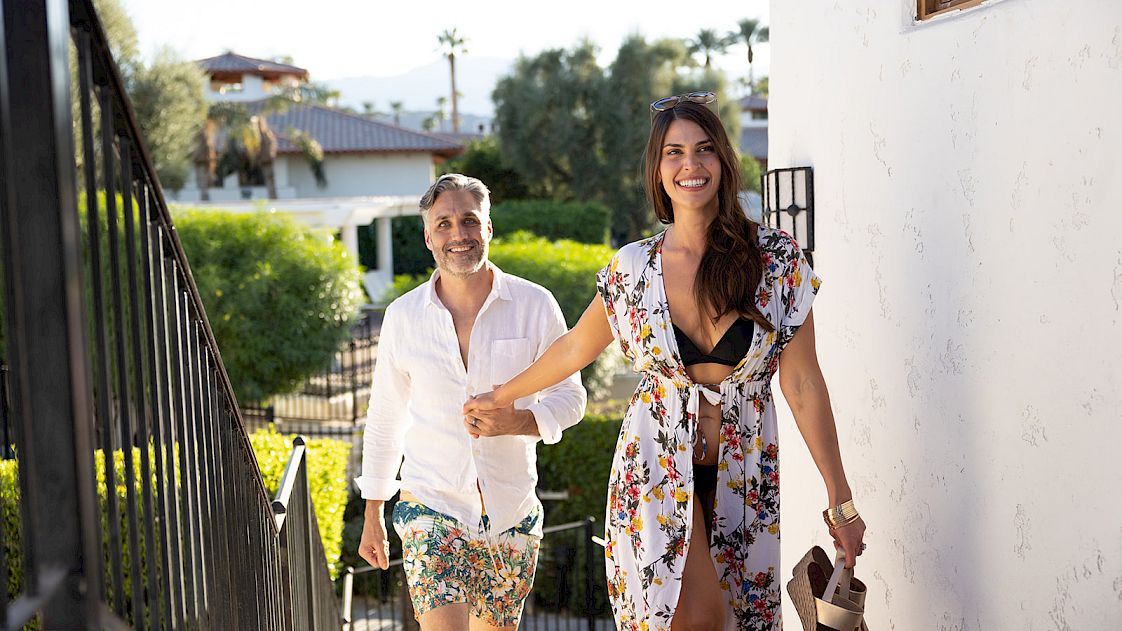 A smiling couple, casually dressed for warm weather, walk up a set of stairs outside of a building. The surrounding area is lush with greenery.